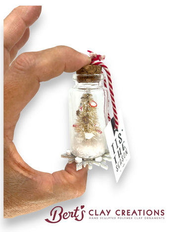 Hand holding a small glass jar with a miniature Christmas tree ornament and a 'Tis the Season' tag, branded 'Bert's Clay Creations'.
